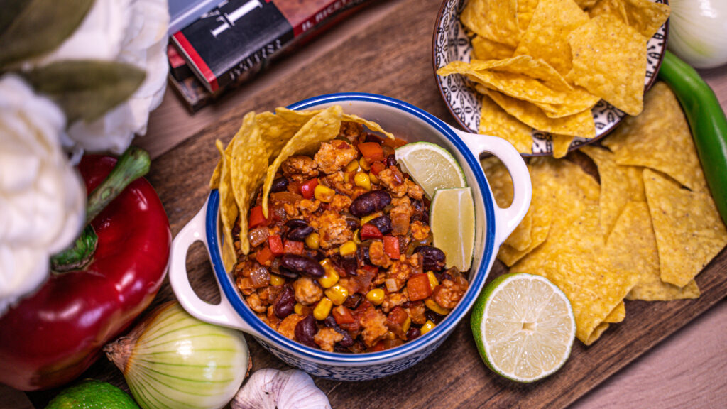 Chilli cu carne de curcan servit într-un bol decorativ, alături de tortilla chips și lime proaspăt.