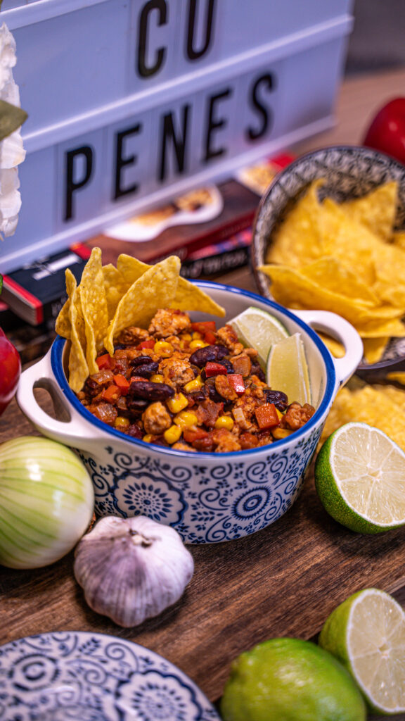 Bol cu Chilli cu carne de curcan, decorat cu tortilla chips și lime, înconjurat de ingrediente proaspete